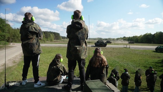 Eine Gruppe Schüler:innen in Tarnkleidung und mit Gehörschutz schaut einem über das Gelände fahrenden Panzer zu. © Screenshot 