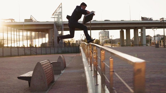 Ein Mann im Anzug und einem Aktenkoffer in der Hand, springt im Sprint über einen Zaun. © Screenshot 