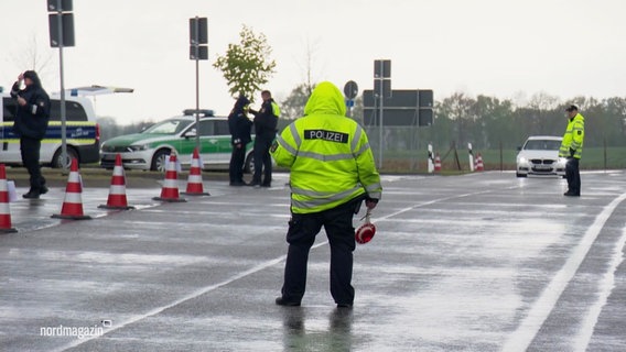 Polizisten haben eine Straße abgesperrt und halten in neongelben Jacken und mit Polizeikellen Autos an. © Screenshot 