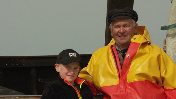 Fischer Uwe Krüger aus Heringsdorf steht in Ölzeug gekleidet neben seinem Enkel Mika. © Screenshot 