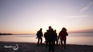 Eine kleine Gruppe Menschen steht bei Morgengrauen mit Foto-Equipment am Strand. © Screenshot 