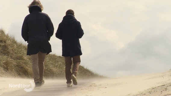 Zwei Menschen in dunklen Anoraks machen einen Spaziergang an einem Strand mit Dünen. © Screenshot 