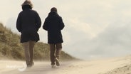 Zwei Menschen in dunklen Anoraks machen einen Spaziergang an einem Strand mit Dünen. © Screenshot 