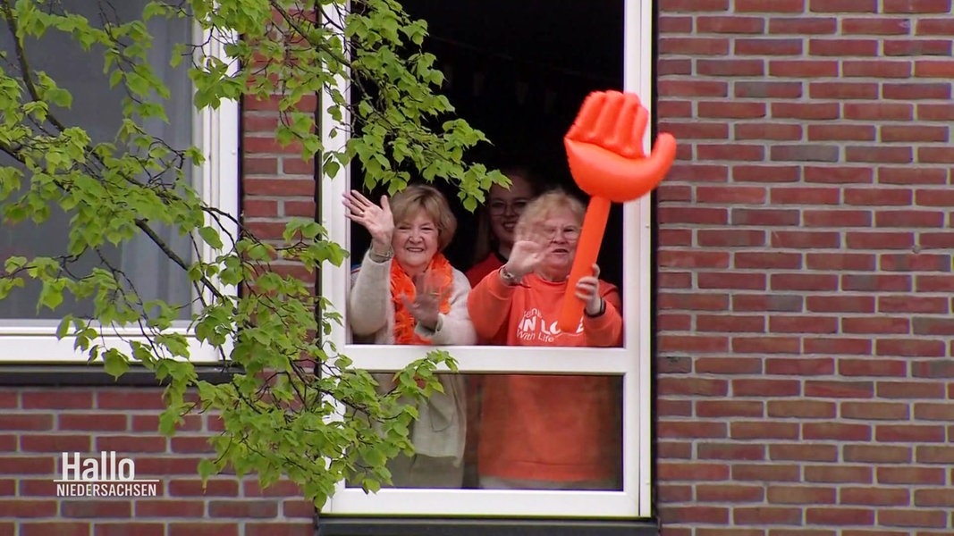Drei Frauen in orangefarbener Kleidung winken mit einer großen aufblasbaren orangenen Hand aus einem Fenster.