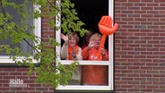 Drie in het oranje geklede vrouwen zwaaien uit het raam met een grote opblaasbare oranje hand.  © Schermafbeelding 