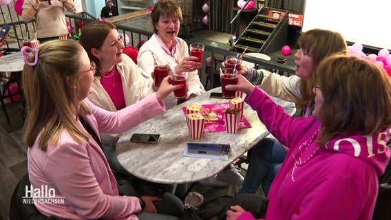 Fünf Frauen in pink-rosa Kleidung sitzen an einem Tisch und stoßen zu Popcorntüten mit ihren Getränken an. © Screenshot 