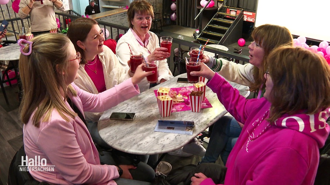 Fünf Frauen in pink-rosa Kleidung sitzen an einem Tisch und stoßen zu Popcorntüten mit ihren Getränken an.