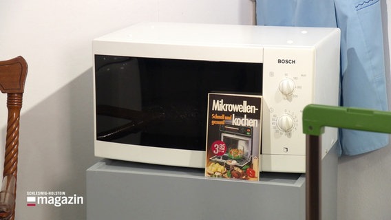 Eine Mikrowelle und ein Kochbuch stehen in einer Ausstellung. © Screenshot 