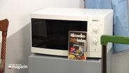 Eine Mikrowelle und ein Kochbuch stehen in einer Ausstellung. © Screenshot 
