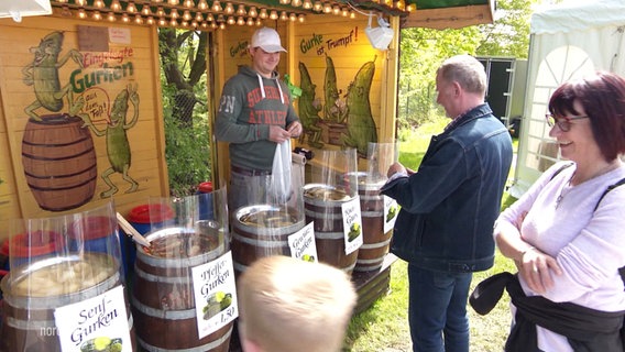 Ein Mann verkauft Gewüzgurken vor seinem Stand stehen zwei Kunden. © Screenshot 