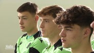 Drei junge Fußballspieler sitzen in hellgrünen Trikots nebeneinander. © Screenshot 