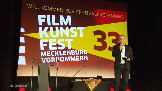 Ein Mann steht auf einer Bühne vor einem großen Plakat mit dem Logo des Filmkunstfestes. © Screenshot 