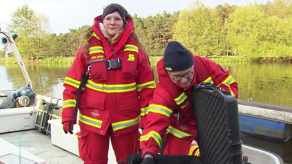 Zwei Beschäftigte der DLRG stehen in Arbeitskleidung auf einem Boot. © Screenshot 