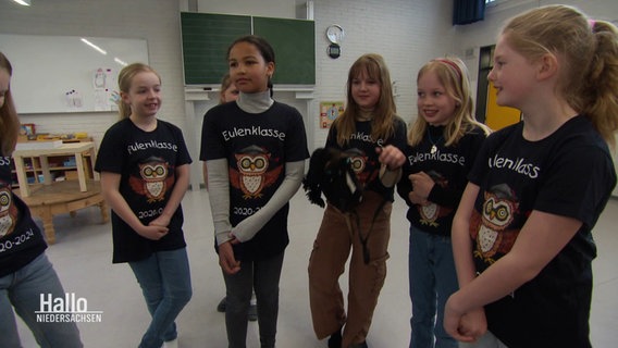 Eine Mädchengruppe mit schwarzen T-Shirts, auf denen "Eulenklasse" zu lesen ist, steht in einem Klasenzimmer. © Screenshot 