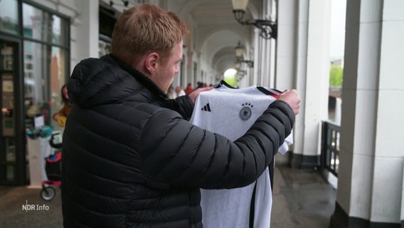 Ein Mann hält ein Trikots der Deutschen Fußballnationalmannschaft in den Händen. © Screenshot 