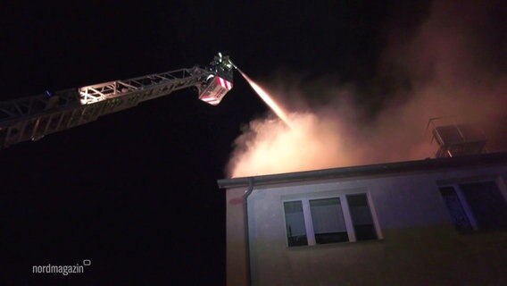 Ein Feuerwehrmann löscht aus einem Leiterwagen heraus ein Feuer im Dach eines Wohnhauses. © Screenshot 