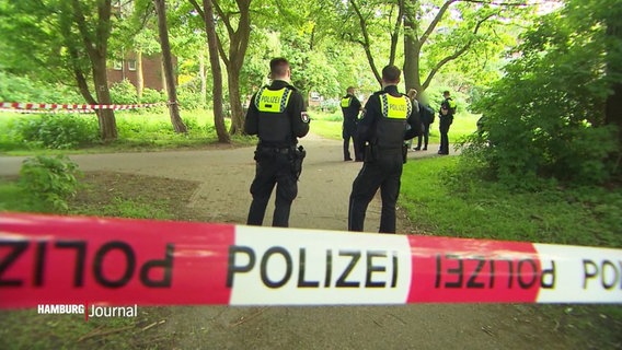 Polizisten stehen in einem Park hinter rot-weißem Absperrband. © Screenshot 