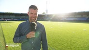 Der Reporter Philip Kahmke steht im leeren Stadion auf dem Fußballfeld. © Screenshot 