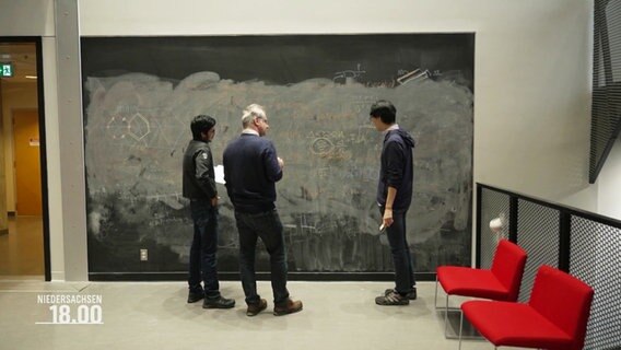 Drei Männer stehen vor einer großen mit Formeln beschriebenen Tafel. © Screenshot 