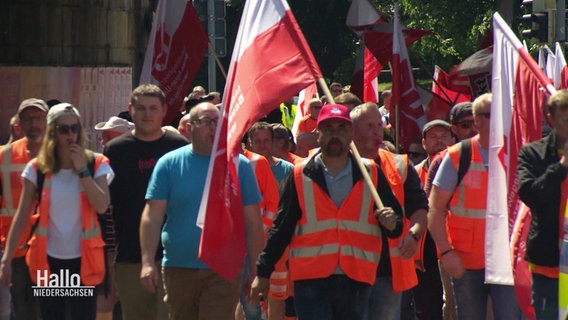 Streikende Bauleute in organgenen Westen schwingen Fahnen und blasen in Trillerpfeifen. © Screenshot 