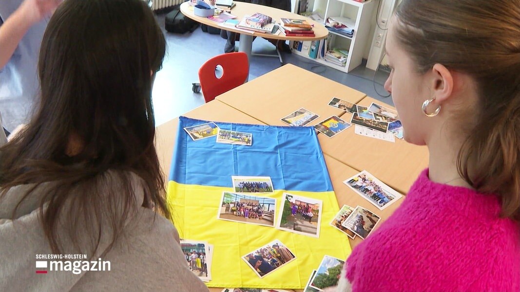 Zwei Schülerinnen betrachten eine ukrainische Flagge und daraufliegende Fotos.