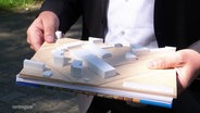 Ein Mann hält ein kleines Holzmodell eines Gebäudekomplexes in den Händen. © Screenshot 