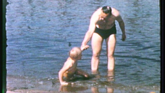 Eine Aufnahme. Ein Mann in Badehose hilft einem blonden Kind im Wasser aufzustehen. © Screenshot 