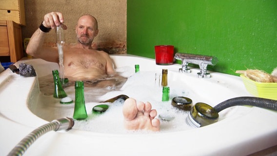 Ein Mann sitzt in einer Badewanne mit Seife. Um ihn herum treiben leere Glasflaschen im Wasser, die er nach und nach so einweicht und von den Etiketten befreit. © Screenshot 