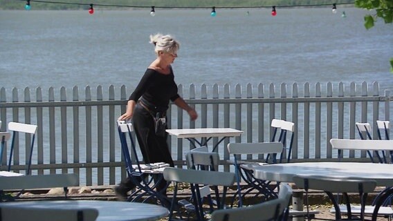 Eine Kellnerin eilt über eine Restaurantterasse. Im Hintergrund ein See. © Screenshot 