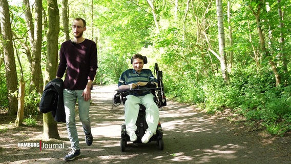 Ein junger Mann läuft neben einem Jungen im Rollstuhl her. © Screenshot 