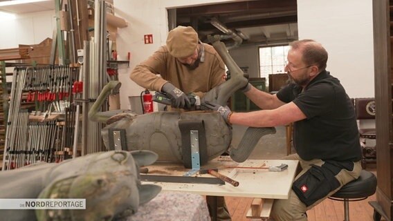 Restaurator Manfred Hübner und sein Mitarbeiter arbeiten in ihrer Werkstatt an der Holzfigur eines Pferdes. © Screenshot 