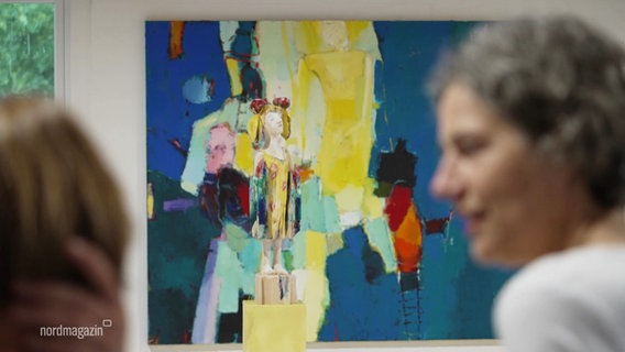 Im Vordergrund unscharf zu erkennen zwei Personen, im Hintergrund zwischen ihnen scharf zu sehen ein farbenfrohes Gemälde. © Screenshot 