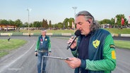 Motorsport-Arena-Sprecher Steffen Schulz mit einem Mikrofon neben der Rennstrecke beim Bundesliga Derby in Güstrow. © Screenshot 