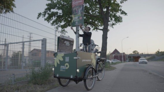 Grünen-Wahlkämpfer Marcel Spittel hängt ein Wahlplakat an einem Laternmast besonders hoch auf. © Screenshot 