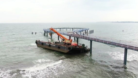 Eine Seebrücke mit Fußgängerweg führt auf Wasser hinaus. Ein schwimmendes Ponton mit Kran wird vom Wasser gegen das Bauwerk gedrückt. © Screenshot 