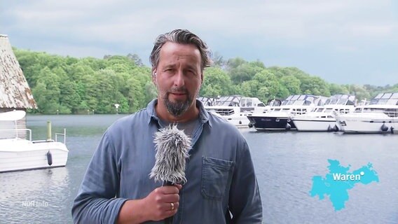 Reporter Stefan Weidig berichtet aus Waren. Er steht vor einer kleinen Bucht in der einige kleine Motoryachten liegen. Über ihm ist der Himmel von grauen Wolken verhangen. © Screenshot 
