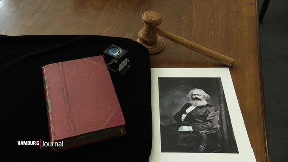 Die Erstausgabe von "Das Kapital" von Karl Marx, daneben eine Lupe, ein Foto des Autors sowie der Hammer einer Auktion liegen auf einem Schreibtisch. © Screenshot 