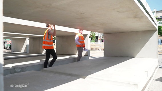 Menschen mit Baustellenschutzkleidung begutachten einen übergroßen Betonrahmen, der in sich Rillen (als Ablaufsteuerung für Wasser) hat. © Screenshot 