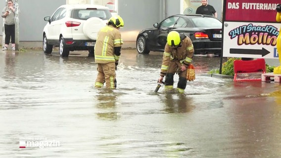 Starkregen: Zwei Mitarbeiter der Feuerwehr stehen auf einer Straße fast knietief im Wasser. © Screenshot 