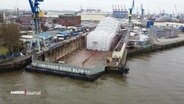Eine eingerüstete Yacht steht auf einem Trockendock. © Screenshot 