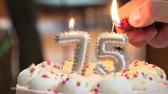Nahaufnahme einer Geburtstagstorte. Darauf zwei Kerzen in der Form einer 75. Die Flamme auf der 7 brennt schon, die auf der 5 wird gerade von einer Hand mit Feuerzeug angezündet. © Screenshot 