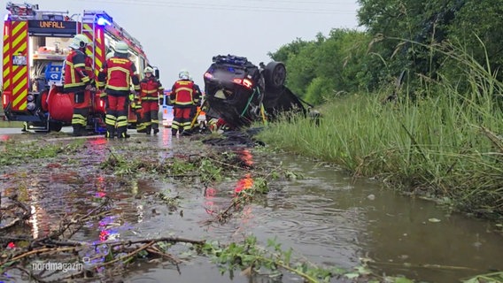 Ein umgedrehtes Auto liegt am Straßenrand, daneben Ein Feuerwehrwagen und mehrere Einsatzkräfte. Im Vordergrund sieht man den stark überschwemmten, schlammigen Grünstreifen. © Screenshot 