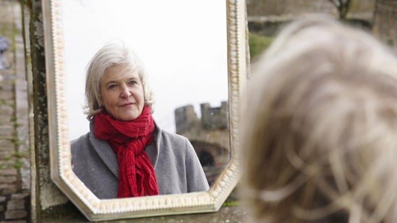Eine ältere Frau mit grauen Haaren blickt sich selbst in einem Spiegel an. © Screenshot 