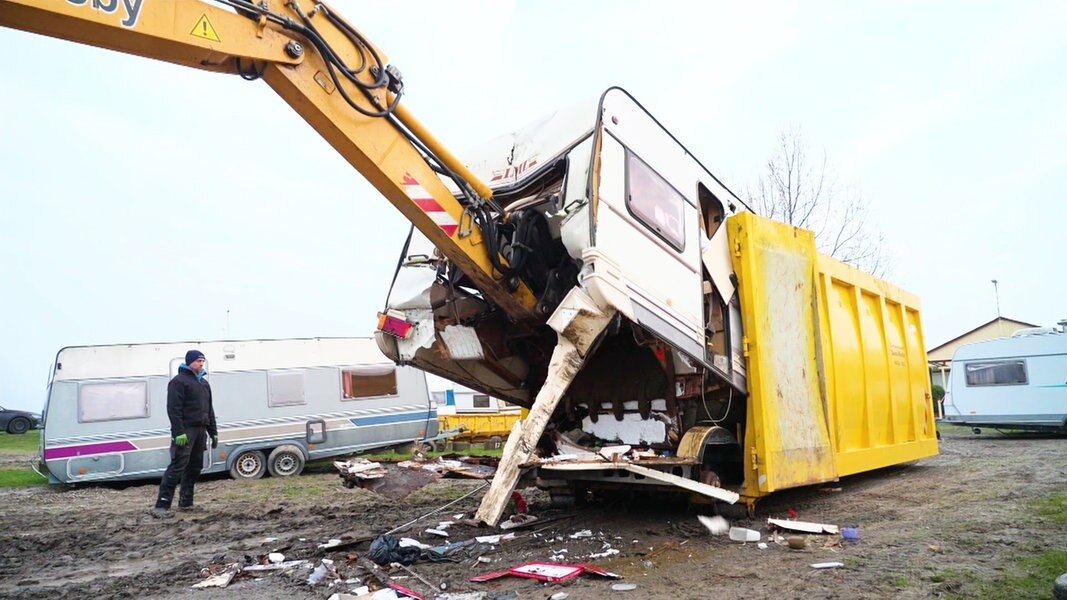 Ein Bagger verfrachtet einen kaputten Camper in einen gelben Müllcontainer.