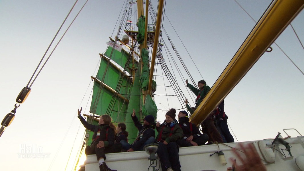 Junge Menschen winken von einem Schiff mit grünen Segeln.