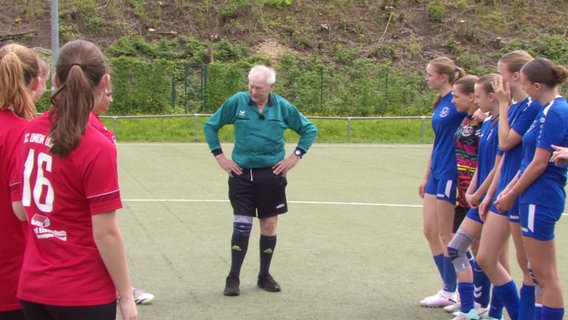 Herbert Schönbeck steht als Schiedsrichter zwischen zwei Jugend-Mädchenteams. Er ist ein Mann über 80, mit grauem und schütterem Haar. © Screenshot 