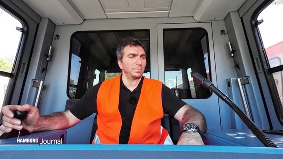 Ein Mann in organgefarbener Warnweste sitzt im Cockpit einer Bahn. © Screenshot 