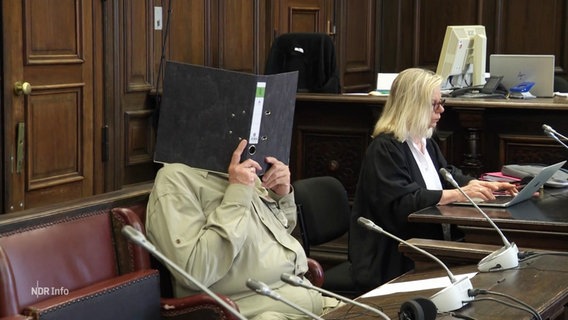 Der Angeklagte hält sich einen Ordner vors Gesicht. © Screenshot 