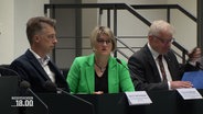 Eine Frau in grünem Blazer und zwei Männer im Anzug. © Screenshot 