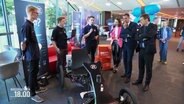 Ein junger Mann mit Mikrofon präsentiert ein Fahrzeug, zwei Männer in Anzügen hören zu. © Screenshot 
